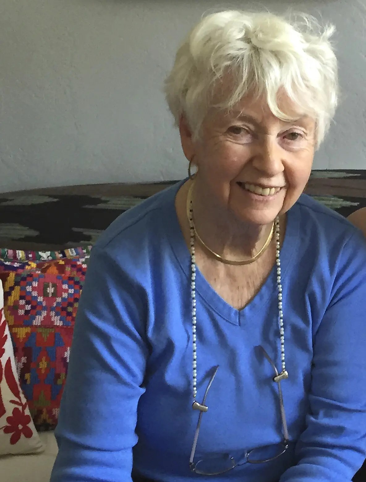 Author Eve Bunting-an older white woman with short white hair, sitting on a couch. She is wearing a long sleeved blue shirt  and a long chain around  her neck. 