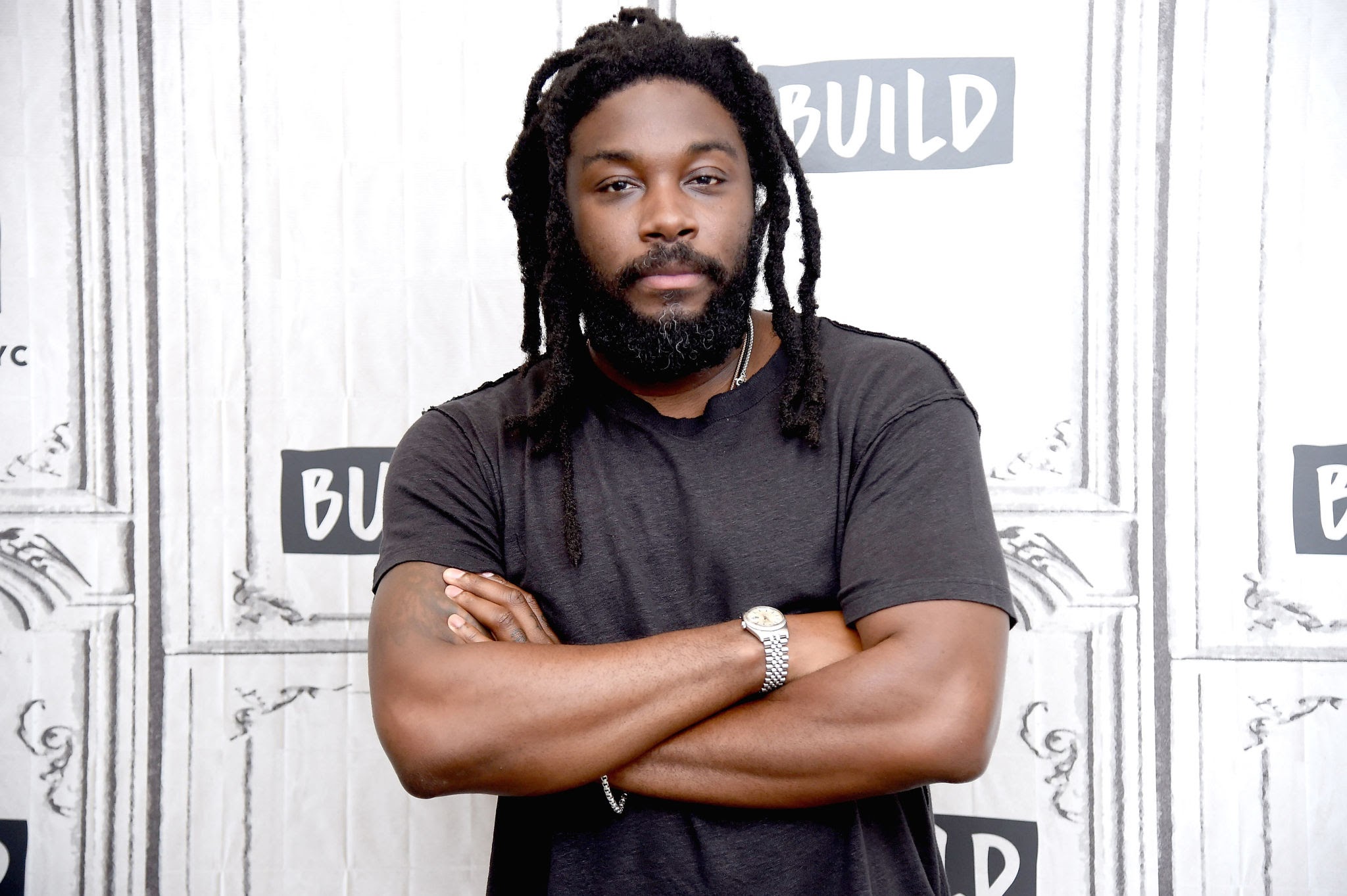 Photo shows a handsome black man with a beard and locked hair. He is wearing a short sleeve black t-shirt and has his arms crossed while looking directly at the camera. He stands againts a predominantly white background with some gray letter text as design elements. 