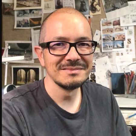 Author Shaun Tan. He is a thin biracial Asian man,  bald, and wears dark rimmed glassed. He  is wearing a faded black t-shirt. He is photographed from chest up in a seated position, and behind him is a bulletin board with several drawings tacked upon it. 
